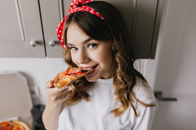 Chica de buen humor comiendo pizza en la cocina. Toma de primer plano de mujer positiva disfrutando de su cena.