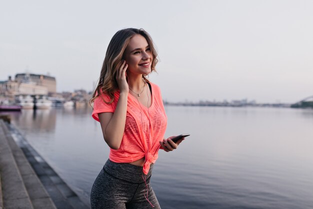 Chica de buen humor escuchando música después del entrenamiento. Corredor femenino atractivo que presenta cerca del río con una sonrisa.