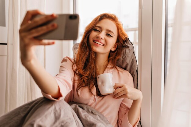 Chica de buen humor haciendo selfie mientras bebe café Hermosa mujer pelirroja disfrutando del té en la cama