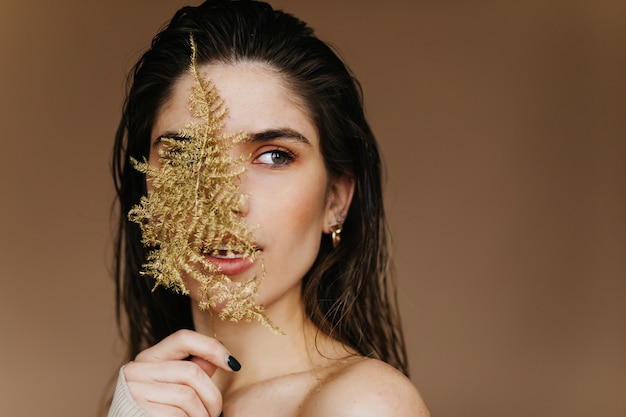 Chica de buen humor lleva joyas posando con plantas. Toma de primer plano de hermosa mujer de pelo negro con hoja verde.