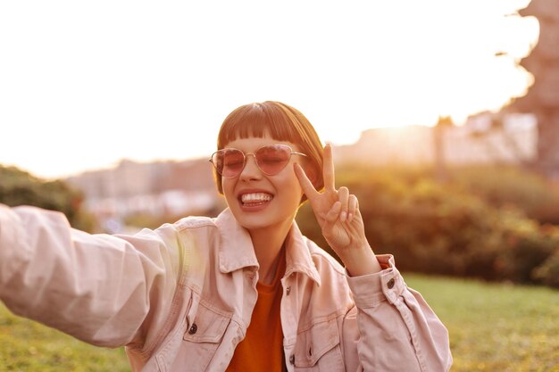 Chica de buen humor se toma selfie durante la puesta de sol Elegante mujer joven con gafas de sol y chaqueta de mezclilla rosa muestra el signo de la paz