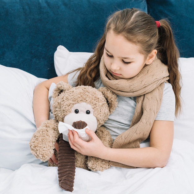 Foto gratuita chica con bufanda alrededor de su cuello cubriendo la boca de un oso de peluche con un pañuelo de papel