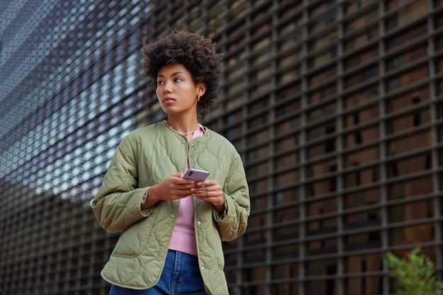 chica con cabello afro rizado usa el teléfono móvil para comunicarse en el tiempo libre al aire libre usa chaqueta y jeans posa al aire libre disfruta del tiempo de recreación mientras pasea por las calles de la ciudad