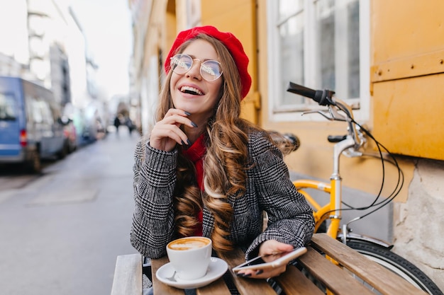 Chica de cabello castaño soñadora riendo mientras toma café en la calle. Retrato de mujer romántica en boina roja sentado en la mesa de madera con una taza de café con leche y bicicleta en el fondo.