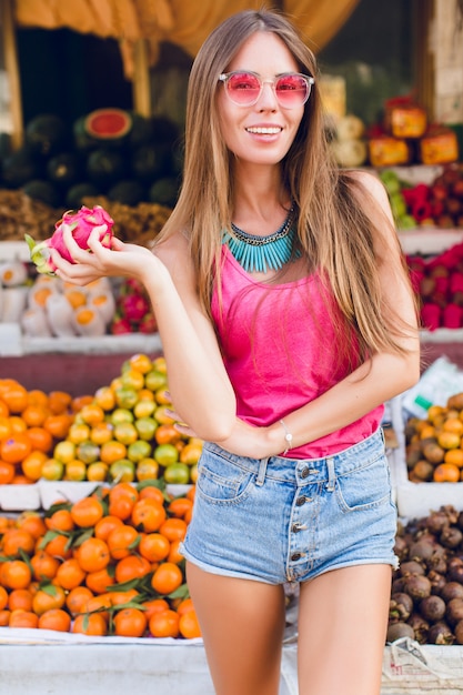 Chica con cabello largo y buen cuerpo en el mercado de frutas tropicales. Lleva gafas de sol rosas, sostiene maracuyá y sonríe
