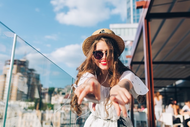 Chica con cabello largo con gafas de sol está escuchando música a través de auriculares en el balcón. Lleva un vestido blanco, lápiz labial rojo y sombrero. Ella está estirando las manos hacia la cámara. Vista desde abajo.