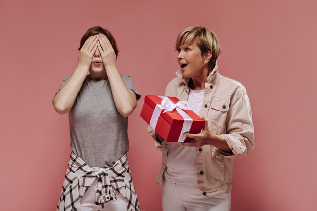 Chica con cabello morena cerrando los ojos con las manos y posando con una mujer rubia en chaqueta beige con caja de regalo roja sobre fondo rosa.