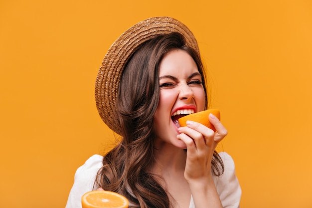 Chica con cabello ondulado en sombrero muerde jugosa naranja sobre fondo aislado.