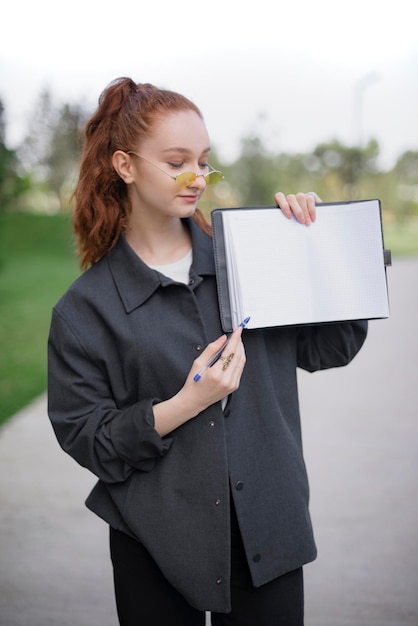 Chica con cabello rojo de pie en el parque mostrando cuaderno abierto