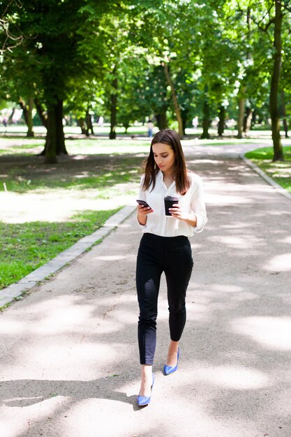 Foto gratuita chica camina con teléfono en la mano y una taza de café en el parque