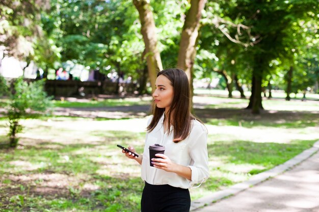 Chica camina con teléfono en la mano y una taza de café en el parque