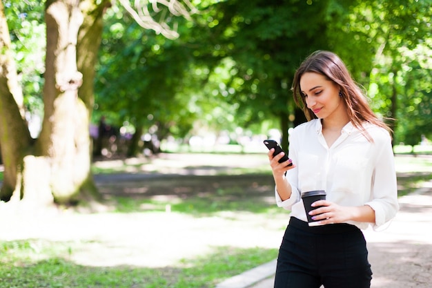 Chica camina con teléfono en la mano y una taza de café en el parque