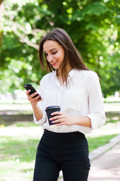 Chica camina con teléfono en la mano y una taza de café en el parque