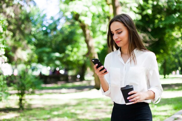 Chica camina con teléfono en la mano y una taza de café en el parque