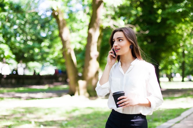Chica camina con teléfono en la mano y una taza de café en el parque