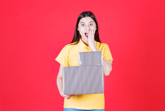 Chica en camisa amarilla con caja de regalo plateada y parece emocionada y sorprendida