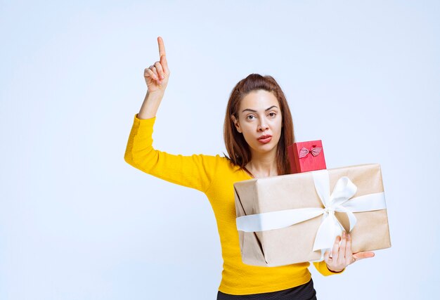 Chica de camisa amarilla sosteniendo cajas de regalo y apuntando hacia arriba.