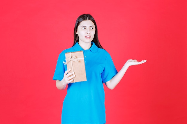 Chica con camisa azul sosteniendo una mini caja de regalo de cartón y llamando a alguien para que se acerque y la lleve