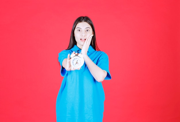 Chica con camisa azul sosteniendo un reloj despertador y dándose cuenta de que llega tarde