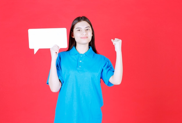 Chica con camisa azul sosteniendo un tablero de información rectangular y mostrando un signo de mano positivo.
