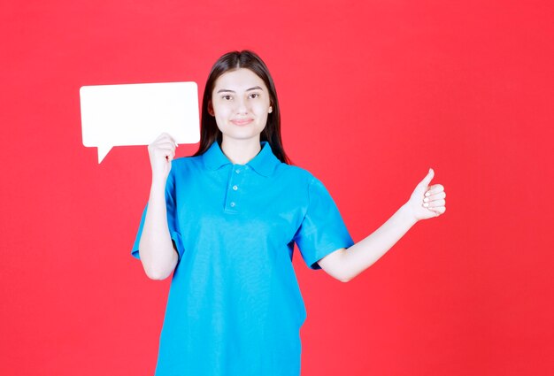 Chica con camisa azul sosteniendo un tablero de información rectangular y mostrando un signo de mano positivo