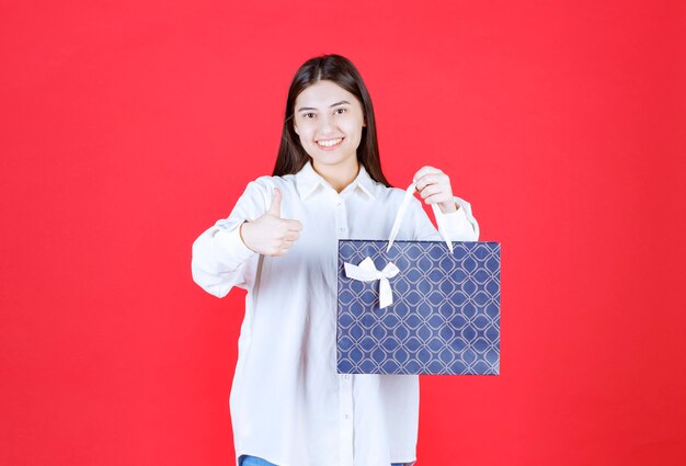 Chica con camisa blanca sosteniendo una bolsa azul y mostrando un signo de mano positivo