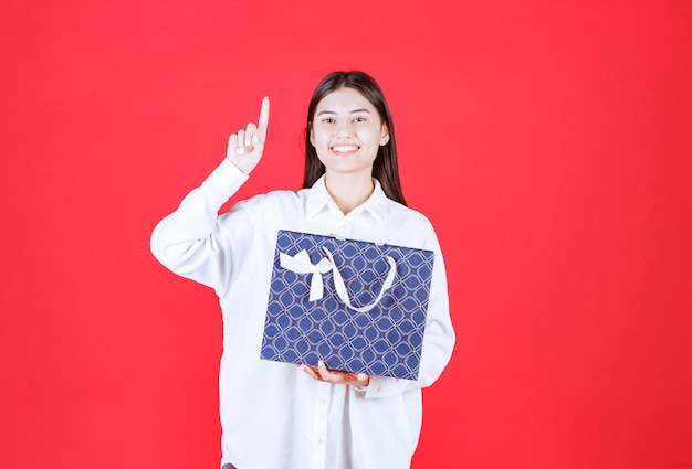 Chica con camisa blanca sosteniendo una bolsa de compras azul y parece confundida y pensativa