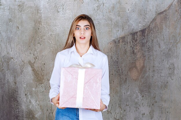 Chica con camisa blanca sosteniendo una caja de regalo rosa envuelta con cinta blanca y parece asustada o aterrorizada