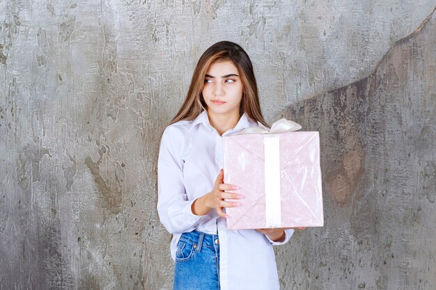 Chica con camisa blanca sosteniendo una caja de regalo rosa envuelta con cinta blanca y parece confundida y vacilante