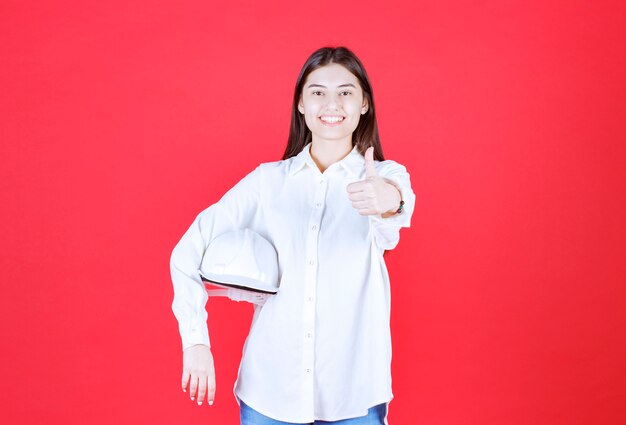 Chica con camisa blanca sosteniendo un casco blanco y mostrando un signo de mano positivo