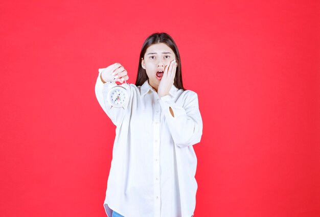 Chica con camisa blanca sosteniendo un despertador y dándose cuenta de que llega tarde