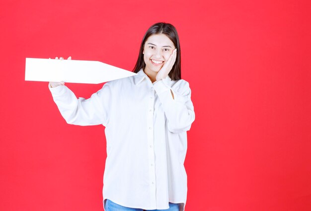 Chica con camisa blanca sosteniendo una flecha apuntando hacia la derecha y parece confundida o pensativa