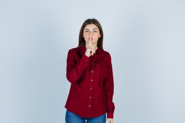 Chica en camisa burdeos mostrando gesto de silencio y mirando misteriosa