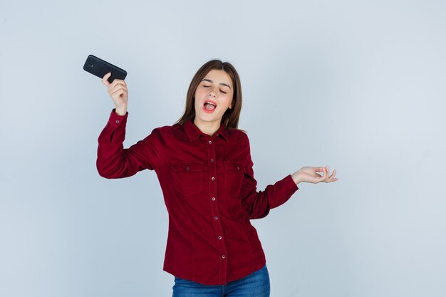 Chica en camisa burdeos sosteniendo un teléfono inteligente, divirtiéndose, disfrutando de la música, bailando y mirando divertido