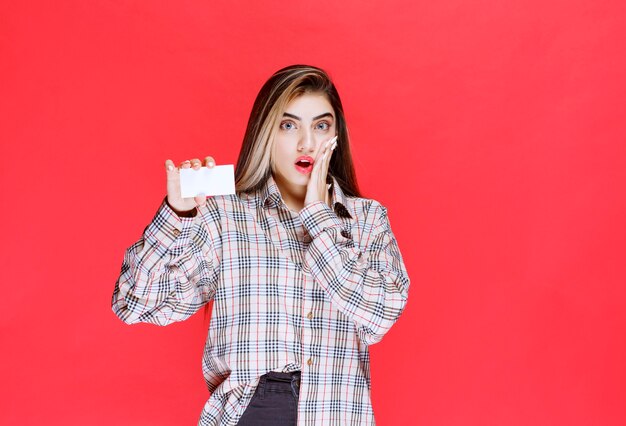 Chica en camisa a cuadros sosteniendo una tarjeta de visita y parece sorprendida