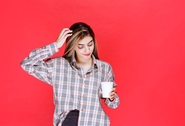 Chica en camisa a cuadros sosteniendo una taza de café desechable blanca y parece pensativa o tiene buenas ideas