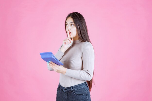 Chica en camisa gris sosteniendo una calculadora azul y mostrando el signo de silencio mientras trabaja