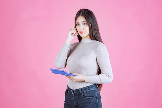 Chica en camisa gris sosteniendo una calculadora azul y pensando en los detalles