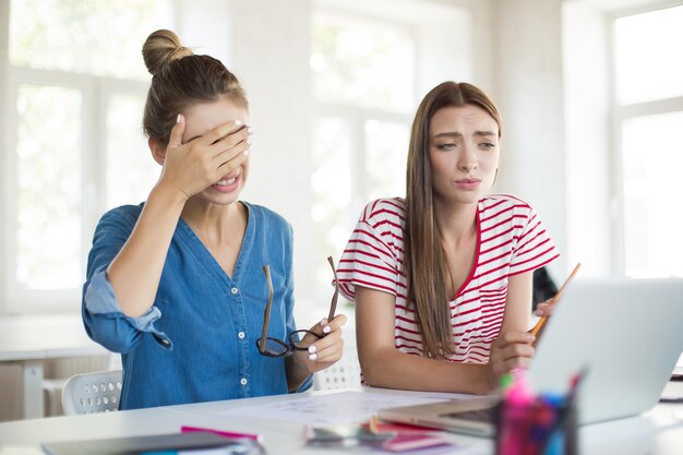 Chica en camisa de mezclilla con anteojos cubriendo tristemente la cara con la mano mientras la chica mira cuidadosamente en la computadora portátil Mujeres jóvenes trabajando juntas en la oficina moderna