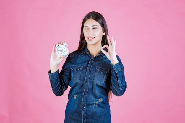 Chica en camisa de mezclilla sosteniendo el despertador y haciendo buena señal