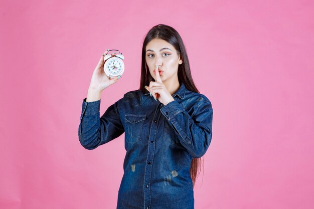 Chica en camisa de mezclilla sosteniendo el despertador y haciendo señal de silencio