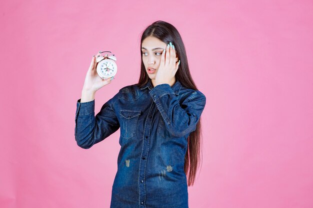 Chica en camisa de mezclilla sosteniendo el despertador y tapándose la oreja debido al anillo