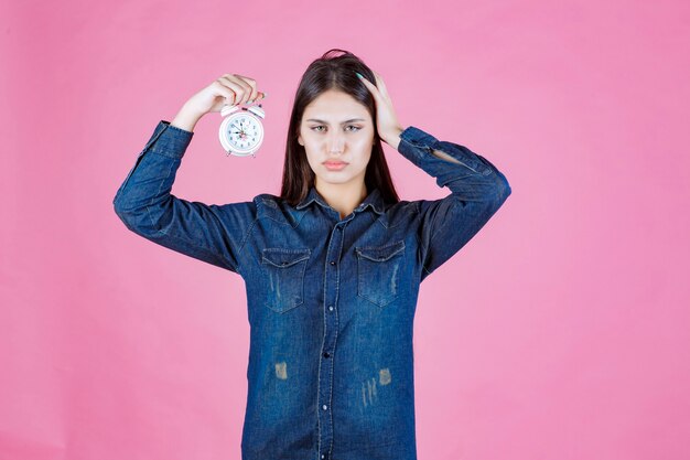 Chica en camisa de mezclilla sosteniendo el despertador y tapándose la oreja debido al anillo