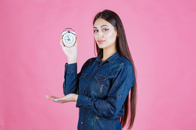 Chica en camisa de mezclilla sosteniendo un reloj despertador y promoviéndolo