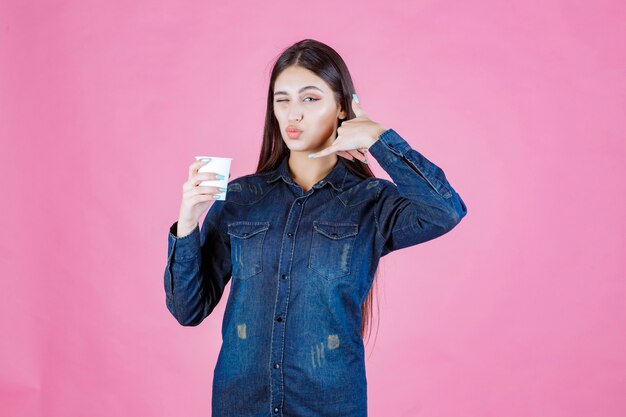 Chica en camisa de mezclilla sosteniendo una taza de café y haciendo señal de llamada