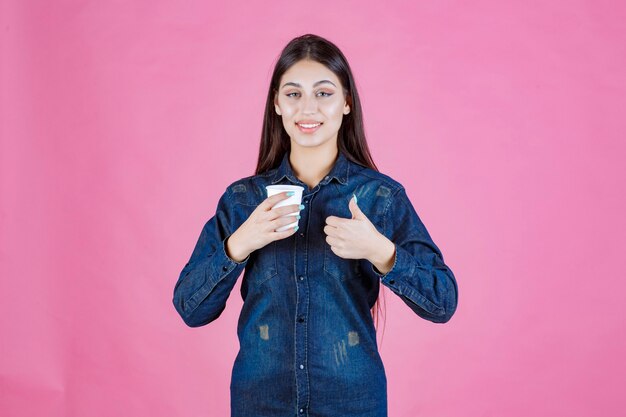 Chica en camisa de mezclilla tomando café y haciendo pulgar arriba