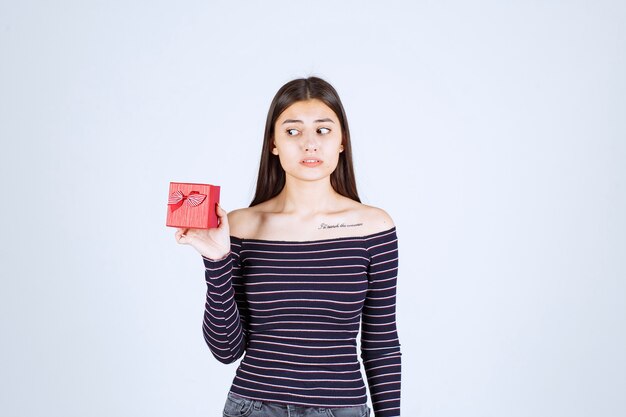 Chica en camisa a rayas sosteniendo una caja de regalo roja, parece confundida y dudosa.