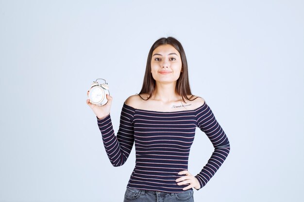 Chica en camisa a rayas sosteniendo un despertador y promocionándolo como un nuevo producto.