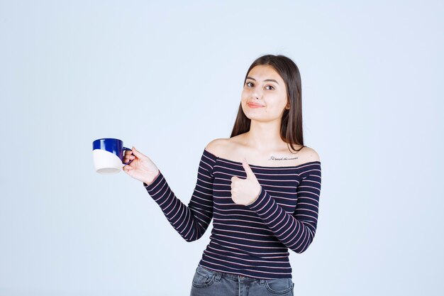 Chica en camisa a rayas sosteniendo una taza de café y mostrando signo de disfrute.