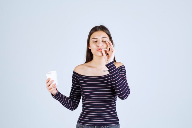 Chica en camisa a rayas sosteniendo una taza de café de plástico y apuntando a un buen gusto.